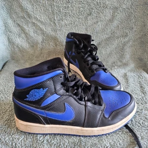 Air Jordan 1 Mid svart/blå - Snygga Air Jordan 1 Mid sneakers från Nike i svart och blått läder med klassisk logga på sidan. Skorna har en vit mellansula och blå yttersula, samt svarta skosnören. Perfekt för dig som gillar streetstyle och ikoniska modeller.