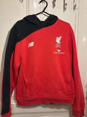 Liverpool FC röd hoodie New Balance (2015-16) - Cool röd hoodie från Liverpool FC med svart axel och huva. Tryck med L.F.C.-logga och Garuda Indonesia på bröstet. Materialet är mjukt och bekvämt, perfekt för dig som gillar sportiga kläder och vill visa ditt stöd för laget.