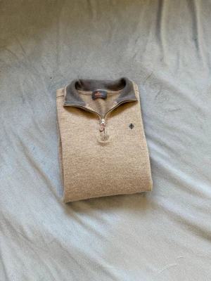 Beige halfzip morris storlek Xs - Snygg beige ziptröja från Morris med grå krage och halv dragkedja. Tröjan har ett diskret broderat emblem på bröstet och är gjord i mjukt material som passar perfekt till lager-på-lager. Klassisk och stilren design.