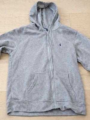 Grå zip-hoodie från Polo Ralph Lauren - Säljer en klassisk grå hoodie med dragkedja från Polo Ralph Lauren. Hoodien har huva med snören, två stora fickor fram och den ikoniska lilla logon broderad på bröstet. Mjuk bomullsblandning och ribbade muddar vid ärmslut och nederkant.