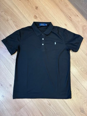 Ralph Lauren pikétröja - Klassisk svart pikétröja från Ralph Lauren med vit broderad logga på bröstet. Tröjan har krage, tre knappar framtill och korta ärmar. Perfekt för en stilren och avslappnad look.