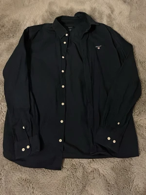 Mörkblå skjorta från GANT - Säljer en klassisk mörkblå skjorta från GANT med vita knappar och diskret logga broderad på bröstet. Skjortan har långärmad design och är tillverkad i bomull, perfekt för en stilren look.