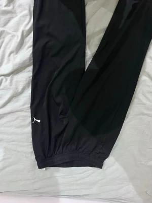 Svarta mjukisbyxor från Puma - Svarta mjukisbyxor från Puma med klassisk logga på vänster ben. Byxorna har resår i bensluten och är tillverkade i ett mjukt och bekvämt material. Perfekta för en avslappnad stil.