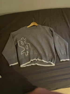 Grå stickad tröja med dollartryck - Cool grå stickad tröja med stort vitt dollartryck på framsidan och texten 'buy happiness' i vitt på ryggen. Tröjan har ribbstickade muddar och nederkant med vit detalj. Perfekt för dig som gillar statement-plagg och streetstyle.