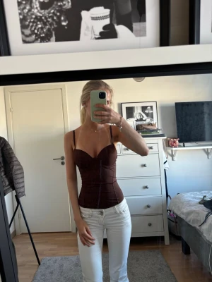 Brun body med smala axelband - Snygg brun body med smala justerbara axelband och markerade kupor. Kroppsnära passform som framhäver midjan och ger en stilren siluett. Materialet ser ut att vara mjukt och stretchigt, perfekt för att skapa en cool look.