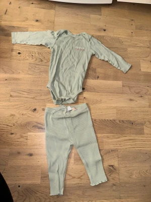 Mintgrönt body och leggings-set - Säljer ett set från GAP med en mintgrön långärmad body och matchande leggings. Bodyn har tryckknappar i grenen och ett diskret tryck på bröstet. Leggingsen är ribbade med liten rosett i midjan och har vågiga kanter vid benslut. Perfekt för en avslappnad och stilren look.
