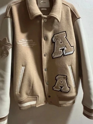 Beige overshirt från Axel Arigato - Snygg beige overshirt från Axel Arigato med vita kontrasterande ärmar och broderade detaljer. Jackan har stora A-applikationer fram och AXEL-text bak, ribbade muddar och tryckknappar. Materialet är ullblandning med inslag av syntet. Perfekt för en trendig look.