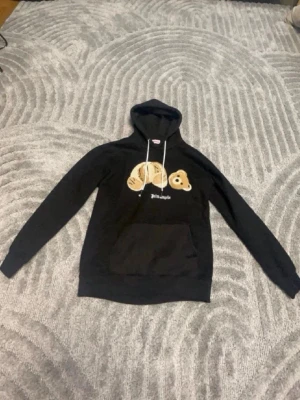 Svart hoodie från Palm Angels med björn - Cool svart hoodie från Palm Angels med ett broderat björnmotiv på bröstet och vit logotyp. Hoodien har huva med vita snören och en stor magficka. Perfekt för dig som gillar streetwear och unika detaljer.