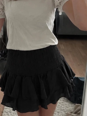 Svart volangkjol med smock - Säljer en svart kortkjol med bred smock i midjan och flera lager volanger. Kjolen har en luftig och flowy känsla som ger en cool look. Perfekt att matcha med en enkel topp för en trendig outfit.