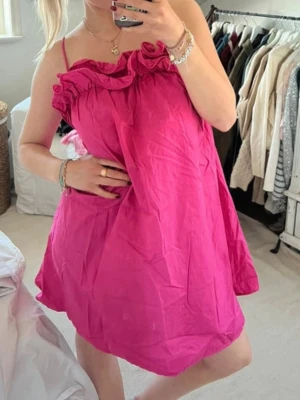 Rosa kort klänning med volang - Supersöt rosa kort klänning med tunna axelband och volangdetalj upptill. Klänningen har en loose passform och är perfekt för varma dagar. Materialet känns lätt och luftigt, vilket gör den bekväm att bära hela sommaren.