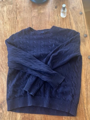 Mörkblå kabelstickad tröja - Säljer en mörkblå kabelstickad tröja med rund hals och ribbade muddar vid ärmslut och nederkant. Tröjan har ett klassiskt flätmönster och är perfekt för att hålla sig varm under kyliga dagar. Passar dig som gillar stilrena och tidlösa plagg.