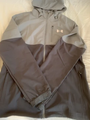 Grå vindjacka från Under Armour - Snygg grå vindjacka från Under Armour med två nyanser av grått, huva och dragkedja framtill. Jackan har logotyp på bröstet, fickor med dragkedja och elastiska muddar med Under Armour-tryck. Perfekt för att hålla vinden borta under utomhusaktiviteter. Haft den i Cirka 1 månad.