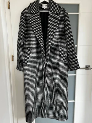 Svartvit hundtandsmönstrad rock NA-KD - Lång rock från NA-KD med klassiskt svartvitt hundtandsmönster. Dubbelknäppt med stora svarta knappar och breda slag. Två snedställda fickor framtill och helfodrad insida. Perfekt för dig som vill ha en stilren och trendig look.