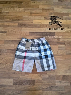 Burberry Shorts - 🩳 Burberry Shorts – Storlek S 🩳 Stilrena Burberry shorts med klassiskt rutmönster, perfekta för sommar, strand eller avslappnad vardag. Elastisk midja med justerbar dragsko ger bekväm passform. Tillverkade i lätt och slitstarkt material med mjuk känsla mot huden. Praktiska fickor för småsaker och Burberry-logga. Liten markering på baksidan. Shortsarna är i nyskick. 📦 Snabb leverans – Kontakta för frågor!