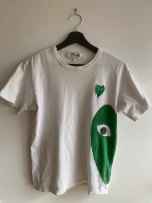 Vit Comme des Garçons Play t-shirt - Vit t-shirt från Comme des Garçons Play med grönt hjärta på bröstet och stort grönt tryck med öga på sidan. Klassisk rund hals och korta ärmar. Tröjan har ett litet hål på baksida nacke men de är inte så märkbart som man ser på bilden. Storleken är S men den sitter mer som xs.