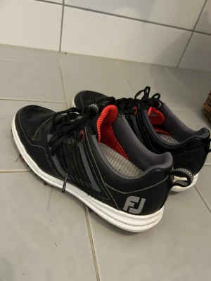 Svarta golfskor från FootJoy - Säljer ett par svarta golfskor från FootJoy med vita och röda detaljer. Skorna har snörning, mesh på insidan och robust yttersula. Perfekta för dig som vill ha bekväma och snygga skor på golfbanan. FJ-logga på sidan och dragflik bak.