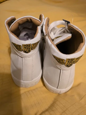Vita high-top sneakers från Gul&Blå  - Snygga vita high-top sneakers från Gul&Blå med broderad G&B-logga och guldfärgade vingar på hälen. Skorna har vita snören, dragkedja på sidan och beige innerfoder. Tiölverkade av skinn. Perfekt för dig som vill sticka ut med coola detaljer och klassisk design.