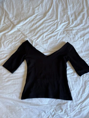 Svart v-ringad topp med trekvartsärm - En svart topp med djup v-ringning och trekvartsärm. Toppens passform är tight och den har en stilren, minimalistisk design som framhäver axlarna. Materialet känns mjukt och stretchigt, perfekt för att skapa en snygg siluett.