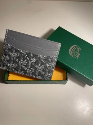 Grå kortfodral från Go4yard - Snyggt grått kortfodral från Go4yard med klassiskt mönster och logotyp på framsidan. Fodralet har flera fack för kort och är tillverkat i lyxigt läder med canvasdetaljer. Perfekt för dig som vill ha en stilren och exklusiv plånbok.