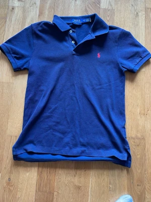 Blå Polo Ralph Lauren t-shirt - Säljer en blå t-shirt från Polo Ralph Lauren med klassisk krage och knappslå. Slim fit-modell med korta ärmar och ikonisk röd logga broderad på bröstet. Materialet är bomull och t-shirten har en stilren och tidlös look.