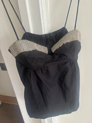 Svart linnetopp med strass från Zara - Snygg svart linnetopp från Zara med tunna axelband och dekorativa strassdetaljer längs överdelen. Toppen har en feminin form med lätt rynkning och är perfekt för dig som vill sticka ut. Materialet känns lätt och bekvämt mot huden.