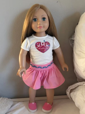 Docka MissMiniMe med kläder  - Säljer en docka från MissMiniMe med långt blont hår, vit t-shirt med hjärtmotiv och rosa kjol. Dockan har rosa skor och flätat bälte. Medföljer original rosa kartong med MissMiniMe-logga. Perfekt för lek och samling.