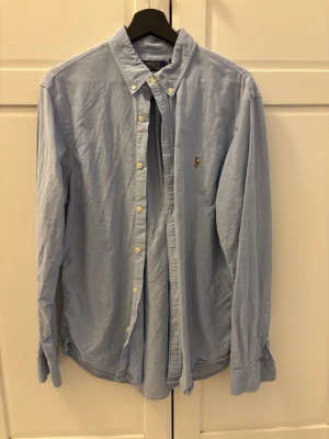 Blå skjorta från Polo Ralph Lauren - Klassisk ljusblå skjorta från Polo Ralph Lauren i slim fit-modell. Skjortan har button-down krage, långa ärmar och knappar framtill. Broderad logga på bröstet. Perfekt för en stilren look.
