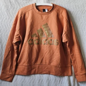 Orange sweatshirt från Adidas - Cool orange sweatshirt från Adidas med stort logotryck framtill. Tröjan har rund halsringning och långa ärmar. Tillverkad i mjuk bomull som känns skön mot huden. Perfekt för en avslappnad och sportig stil.