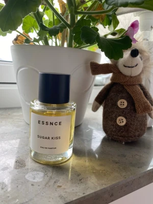 ESSNCE Sugar Kiss Eau de Parfum - ESSNCE Sugar Kiss Eau de Parfum i en stilren, rund glasflaska med mörkblått lock. Parfymen har en ljusgul vätska och etiketten är enkel och modern. Perfekt för dig som gillar söta och fräscha dofter. Mängd enl bilder