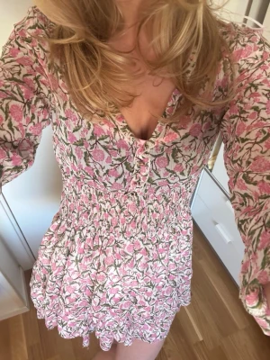 Blommig kortklänning med volang - Söt kortklänning med blommigt mönster i rosa och grönt på vit botten. Klänningen har v-ringning, knappar framtill, smockad midja och volangdetaljer längst ner. Långärmad och luftig, perfekt för vår och sommar.