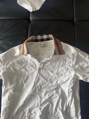 Vit Burberry piké med rutigt krage - Säljer en vit pikétröja från Burberry med den klassiska beige, svarta och röda rutiga kragen. Tröjan har korta ärmar och knappar vid halsen. Materialet är mjukt och bekvämt, perfekt för en stilren look. L men är mer som M/S