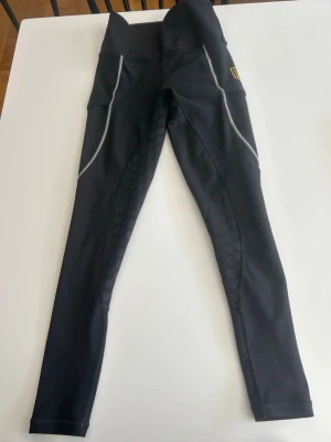 Svarta tights från Maya Delorez - Säljer ett par svarta sporttights från Maya Delorez med hög midja och snygga grå detaljer längs benen. Tightsen har mönstrade paneler med logotyptryck och en gul logotyp på sidan. Perfekta för träning och aktivitet.