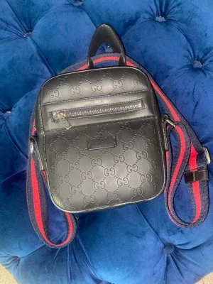 Svart axelväska från Gucci - Snygg svart axelväska från Gucci i läder med präglat GG-mönster över hela väskan. Väskan har en framficka med dragkedja och en justerbar axelrem i blått och rött. Perfekt för dig som vill ha en trendig och exklusiv accessoar.