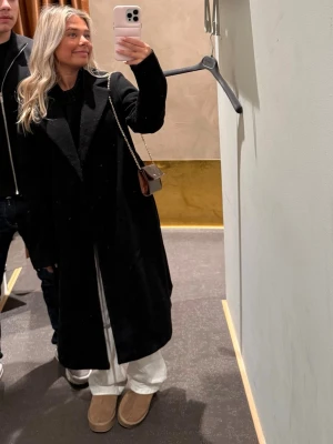 Svart lång kappa med klassisk krage - Säljer en svart lång kappa med klassisk krage och rak passform. Kappan har diskreta fickor och är tillverkad i ett tjockt material som passar bra under kyliga dagar. Perfekt för dig som vill ha en stilren och tidlös look.