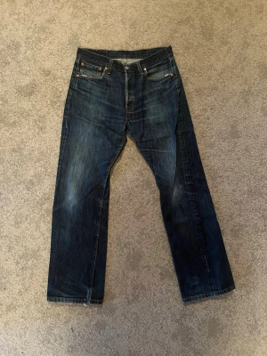 Levi's 501 - Säljer ett par klassiska mörkblå jeans från Levi's med raka ben och femficksdesign. Jeansen har slitna detaljer vid benslut och en snygg tvätt med ljusare partier. Storlek W33 L32. Hör av er vid fler funderingar✅