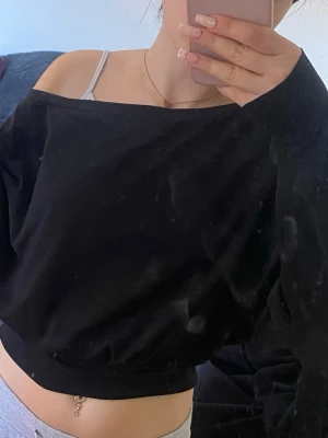 Svart offshoulder croppad topp - Svart croppad topp med offshoulder-design och långärmade ärmar. Toppens material känns mjukt och stretchigt, perfekt för att skapa en avslappnad och trendig look. Passformen är loose och den har bred mudd nedtill som ger en snygg siluett.