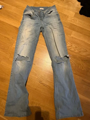 Ljusblå slitna jeans med hål - Ljusblå lågmidjade jeans med bootcut och slitna detaljer på knäna. Jeansen har klassisk femficksdesign och tydliga hål på båda knäna för en trendig look. Perfekta för dig som gillar en avslappnad och cool stil.