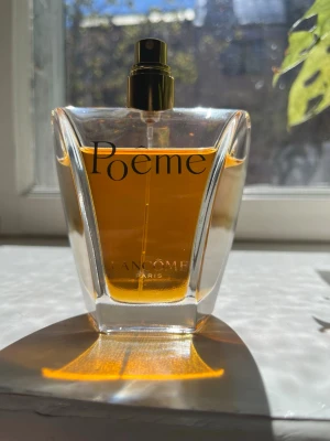 Lancôme Poême Eau de Parfum 100ml - Lancôme Poême Eau de Parfum i en elegant glasflaska med guldtonad spray och tydlig svart text. Flaskan har en unik, geometrisk form och innehåller en djup bärnstensfärgad parfym. En klassisk och ikonisk doft från Lancôme som utstrålar lyx. 100ml