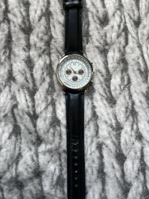 Klassisk svart klocka med läderband - Stilren klocka med svart läderband och rund silverfärgad boett. Urtavlan är vit med tre mindre subdials och tydliga siffror. Perfekt accessoar för dig som gillar en tidlös look.