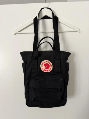 Svart Fjällräven Kånken ryggsäck - En klassisk svart Kånken ryggsäck från Fjällräven med det ikoniska runda röda och vita logotypmärket på framsidan. Ryggsäcken har två axelband, handtag upptill och ett stort huvudfack med dragkedja samt en mindre frontficka. Materialet är slitstark syntet.