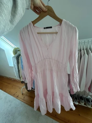 Ljusrosa kortklänning med v-ringning - Söt ljusrosa kortklänning från Zara med v-ringad hals och trekvartsärm. Klänningen har smock i midjan och volang nedtill för en feminin look. Perfekt för varma dagar och enkel att styla med sneakers eller sandaler.