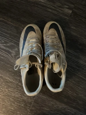 Nike Mercurial vita fotbollsskor - Säljer ett par Nike Mercurial fotbollsskor i vitt med svart swoosh och mönster. Skorna har klassisk snörning och robusta dobbar för bra grepp på planen. Materialet är syntetiskt och designen är smidig och lätt för snabbhet och kontroll.