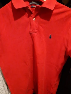 Röd polo från Ralph Lauren Slim Fit - Säljer en klassisk röd polo från Ralph Lauren i slim fit-modell. Tröjan har krage, knappslå och den ikoniska blå logon broderad på bröstet. Materialet är bomull och passformen är tight, perfekt för dig som gillar stilrena och snygga toppar.