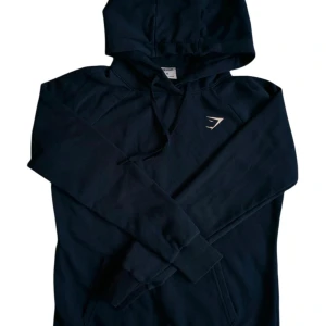 Mörkblå hoodie från Gymshark - Säljer en mörkblå hoodie från Gymshark med klassisk huva och dragsnören. Hoodien har ett diskret vitt Gymshark-logotryck på bröstet och ribbade muddar vid ärmslut och nederkant. Perfekt för en avslappnad och sportig look. Tröjan är i nyskick säljer den för den är för liten för mig