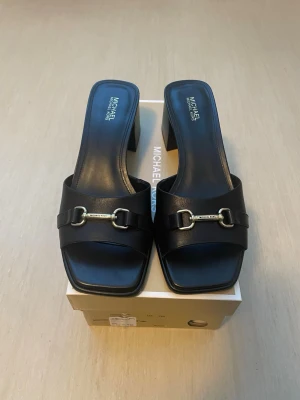 *NY*Svarta Block Heel Carson sandaler från Michael Kors stl 7M (37) - Svarta Lädersandaler med silverdetaljer. Nya i kartong med prislapp, stl 7=37, normal i stl.   Ord pris: 2500 sek, Outlet: 1660 sek,  Pris nu: 700 sek   100% of the leather in this product comes from tanneries receiving Leather Working Group Gold- or Silver-certification in recognition of their environmental performance. • Mule • Leather • Lining: polyurethane blend • Sole: rubber • Open toe • Slip on • Heel height: 1.5” • Heel type: stacked 