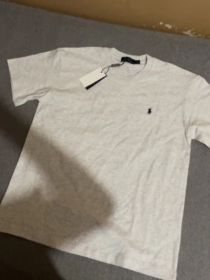 Grå t-shirt från Polo Ralph Lauren - Snygg grå t-shirt från Polo Ralph Lauren med klassisk rund hals och korta ärmar. T-shirten har en liten mörkblå broderad logga på bröstet och är gjord i mjuk bomull. Perfekt basplagg till garderoben.