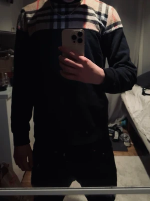 Svart sweatshirt med Burberry-rutor - Säljer en svart sweatshirt från Burberry med klassiskt rutmönster i beige, rött och svart över axlar och bröst. Tröjan har rund halsringning och långa ärmar. Materialet känns mjukt och bekvämt, perfekt för en stilren och trendig look.