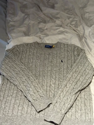 Grå kabelstickad tröja Polo Ralph Lauren - Säljer en grå kabelstickad tröja från Polo Ralph Lauren med rund hals och diskret logga broderad på bröstet. Tröjan har ribbade muddar och är tillverkad i mjukt material som passar perfekt när det är kyligt ute.