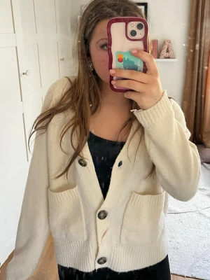 Krämvit kofta med stora knappar - Mysig krämvit kofta med stora mörka knappar och två rejäla fickor framtill. Den är helt som ny.