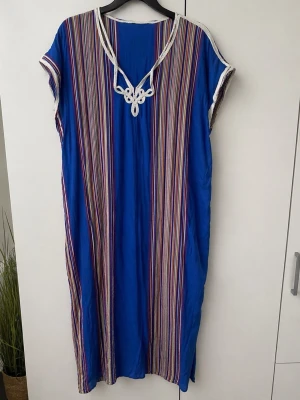     Blå kaftan från Marocko – strl XXS–S - Fin blå kaftan/klänning med färgglada ränder och vita detaljer, direkt från Marocko. Unik och livfull design som ger en härlig, somrig känsla. Lös och luftig modell som är både bekväm och stilren – perfekt för semester, strand eller vardag.  Storlek: passar XXS–S.  Lätt material som faller fint.  Fint skick.